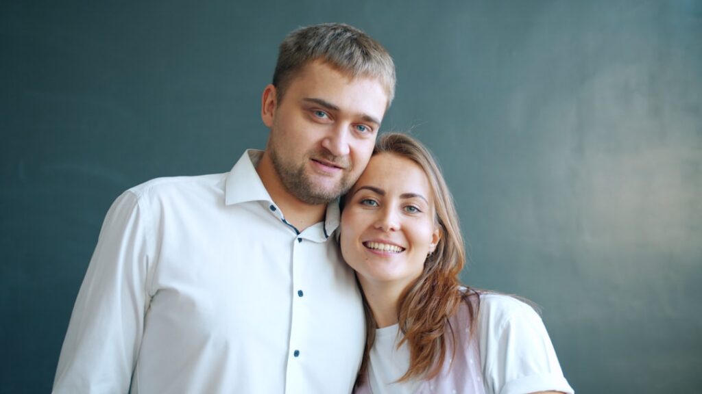 Beautiful couple in love man and woman smiling standing together on gray background, girl putting her head on guy's shoulder. Youth and relationship concept.