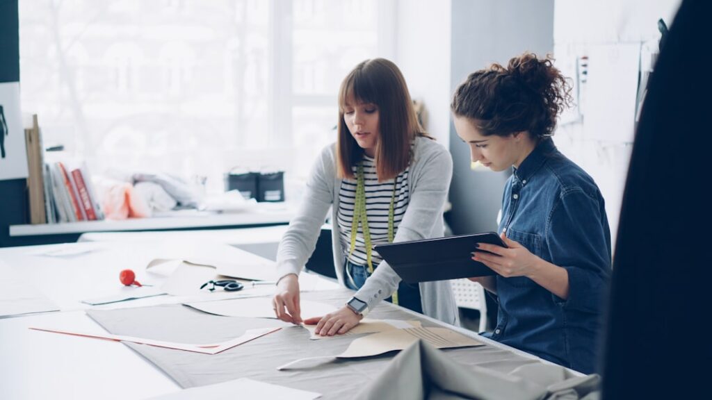 Attractive female coworkers of sewing studio are working with patterns and textile, outlining cutout, talking and using tablet. Friendly cooperation and communication concept.