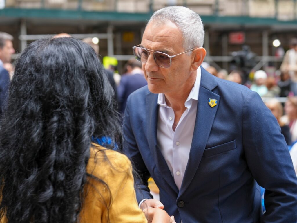 A Ukrainian rally unfolds in downtown Manhattan, where community members, NYC officials, and uniformed police gather in solidarity. A woman draped in a blue and yellow flag stands solemnly among the crowd, while a young attendee holds a folded Ukrainian flag during a formal civic ceremony. The event reflects unity, patriotism, and public support for Ukraine in the heart of New York City. Captured by Olek Buzunov @buzunov.photoart, documenting moments of resilience and civic engagement.